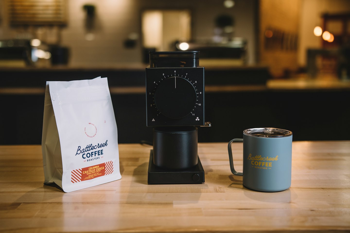 Sacchetto di caffè bianco, macinacaffè e tazza verde acqua su un bancone di legno.